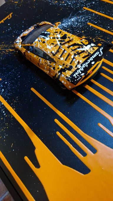 Ein orangefarbenes Modellauto auf einer schwarz-orangefarbenen gespritzten Unterlage.