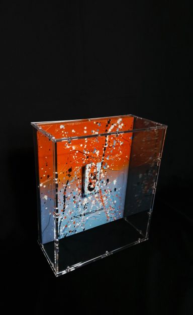 Durchsichtige Wahlurne mit orange-blauem Design und Schlitz an der Oberseite.